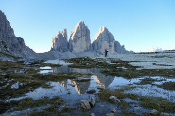 Obraz premium A woman enjoying the view on the Tre Cime di Lavaredo (Drei Zinnen), mountains in Italian Dolomites. The mountains are reflecting in small paddle. Desolated and raw landscape. Early morning. Daybreak