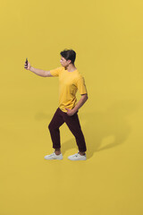 Fototapeta na wymiar Taking selfie on the go. High angle view of young man on yellow background. Boy in motion. Human emotions and facial expressions concept. Full length portait, copyspace for ad. Fashion, retro style.