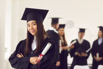 Portrait of a confident and proud Asian female student in a university graduate gown and with a diploma in her hands. Woman posing with crossed arms in classroom on the background of classmates.