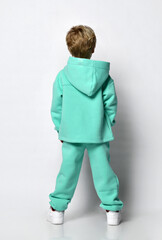 full-length rear view of a little preschooler or schoolboy in a warm turquoise tracksuit with a hood and white sneakers. Isolated over gray background with copy space. Sports