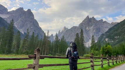 Fototapeta na wymiar A man with a big hiking backpack enjoying the distant view on Italian Dolomites. There is a lush green meadow with a wooden fence around it. Morning fog in the valley. Thick forest on the slopes