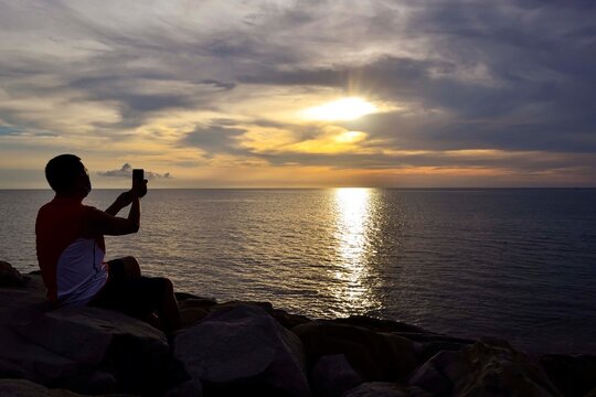 A man wearing face mask uses a smartphone to take pictures of beautiful sunset in Tanjung Aru Kota Kinabalu beach in Borneo Sabah Malaysia