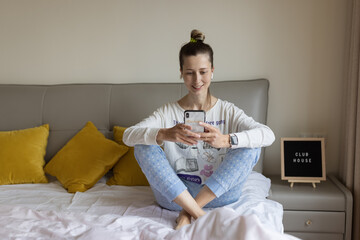 Young caucasian woman using mobile phone and wireless headphones, resting, sitting on bed at home...