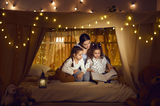 Mother Telling Bedtime Story To Children. Mommy And Little Daughters Sitting In Magic Blanket Fort In Cozy Dark Playroom, Reading Book Of Good Fairy Tales And Enjoying Quiet Family Evening At Home