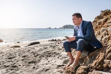 Latin man in elegant suit at the beach using smartphone sitting on the rocks. Escape from city chaos searching a peaceful place.