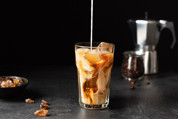 Pouring milk into a glass with iced coffee over black background.
