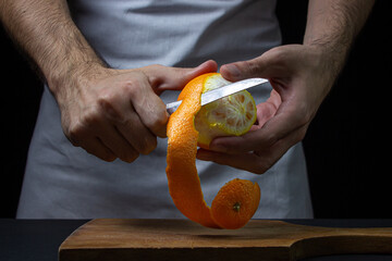 Orange on a dark background. The chef is peeling an orange. Male hands cut orange peel on black background