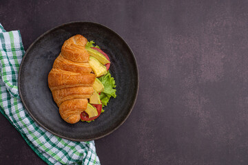 Top view of a croissant with avocados sliced, tomato, and lettuce on a black plate placed on a dark gray background. Space for text. Concept of foods