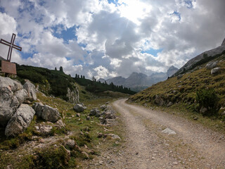 Fototapeta na wymiar A gravelled road leading through Italian Dolomites. There is a metal cross on the boulder next to the road. High mountains around. The slopes are green. Overcast. Spirituality and calmness.