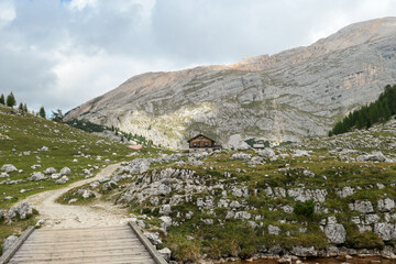Fototapeta na wymiar A gravelled road leading through a small wooden bridge to a cottage in Italian Dolomites. High and sharp mountains behind the cottage. Meadow with many boulders in front. Remote and desolate place.
