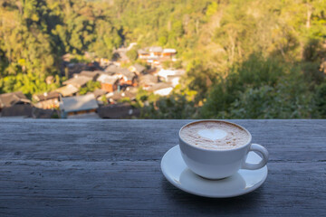 A cup of coffee on wooden table with rural village and mountain view background.