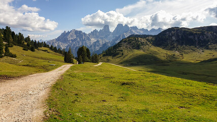 A wide gravelled road in Italian Dolomites from the top of Strudelkopf leading towards the valley. A panoramic view on the high mountains. Sunny day. A few clouds above the peaks. Lush green plateau