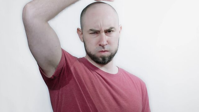 A bald, bearded man raises his hand and shows a spot on his t-shirt from a wet armpit. White background. The concept of sweating and hyperhidrosis