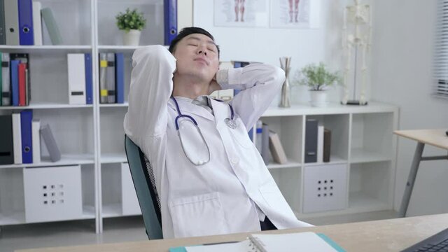 chinese doctor relaxing on the office chair with hands behind head is getting back to work after exercising his neck and stretching his arms.