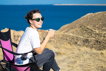 Young woman traveler in sunglasses holding iron mug cup with tea or coffee, enjoying morning with sea scenery in nature landscape, sitting in camp.Travel camping and adventure lifestyle concept.