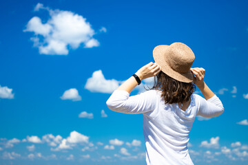 Back view of brunette woman in summer hat and white cloth looking into the distance on blue sky with clouds background. Summer travel and adventure concept. Enjoying life, freedom. Copy space