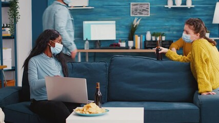Cheerful woman holding beer bottle looking at laptop discussing with african friend in living room wearing face mask to prevent coronavirus spread in time of global pandemic. Conceptual footage.