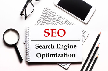 On a white background glasses, a magnifier, pencils, a smartphone and a notebook with the text SEO...