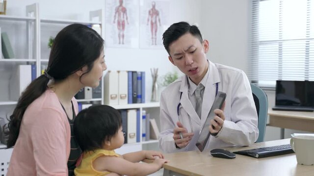 asian pediatrician is tapping the screen with a pen while explaining the test results to the mother and her kid on a touchpad in his clinic office.
