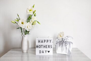 Gift box trendy wrapped in silk fabric in Furoshiki technique, light box with lettering Happy Mother's Day, white flowers Eustoma or Lisianthus in vase on wooden background. Zero Waste Life Concept