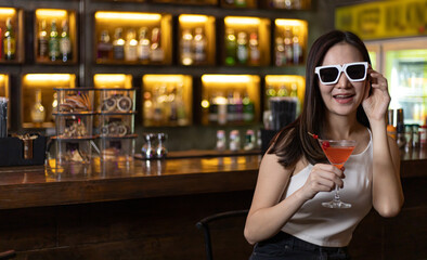 Young Asian woman with black glasses enjoys drinking a cocktail in front of a vintage bar, Relaxing activities after work or hangouts, Place of entertainment for young adolescents or night club party.