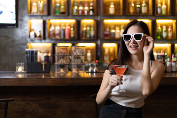 Young Asian woman with black glasses enjoys drinking a cocktail in front of a vintage bar, Relaxing activities after work or hangouts, Place of entertainment for young adolescents or night club party.