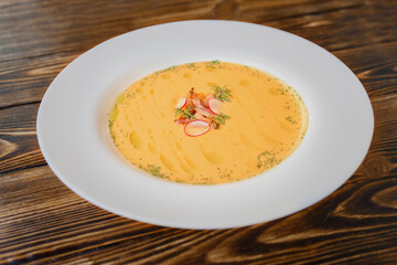 Pumpkin cream soup garnished with slices of bacon and radish in a white plate on a wooden table.