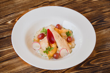 Chicken breast salad with melon on white plate and wooden background, healthy for people who control their weight. Chicken dish garnished with grapes, radish slices and herbs.