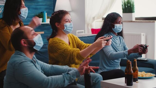Excited multiracial gamer women sitting on couch playing video games on console during new normal party wearing protective masks. Friends using wireless controllers keeping social distancing