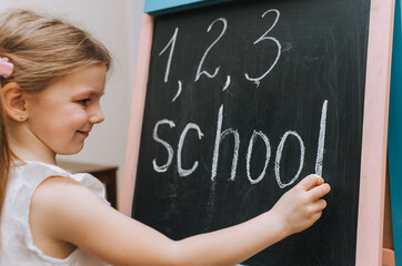 The red-haired little girl, a schoolgirl, writes diligently on a wooden black board, an easel, holding white chalk, the word school and different numbers in her hand. Photography, copy space, concept.