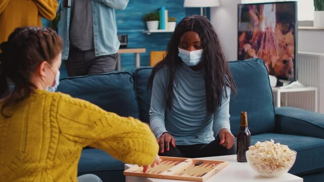 Group of cheerful multiethnic friends playing board games backgammon board games keeping social distancing wearing face mask to prevent infection with covid19 during global pandemic drinking beer