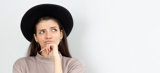 Beautiful female with puzzled expression, keeps finger on lips, looks aside with bewilderment,...