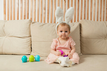 little girl in a headband with bunny ears with egg toys sits in a beige sofa with a basket with a cotton flower