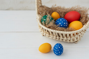 Colored eggs lie in basket on fluffy dried grass, two eggs are near on white table, copy space for text. Preparations for easter holidays.