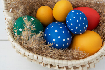 Close up image of colored eggs lie in basket on fluffy dried grass, two eggs are near on white table, copy space for text. Preparations for easter holidays.