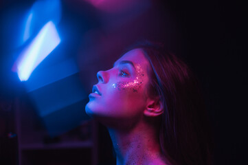 Blurred night art portrait of a woman in neon. Portrait of a skinny brunette with glitter on her face in purple and blue light, looking to the side and glare - obrazy, fototapety, plakaty