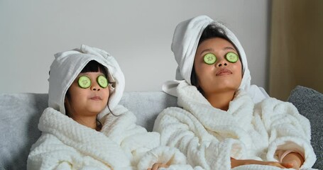 Portrait of asian mother with ethnic daughter girl child wear white bathrobes and towels lies on sofa doing cosmetic procedures natural anti-wrinkle mask to moisturize skin put cucumber pieces on eyes - Powered by Adobe