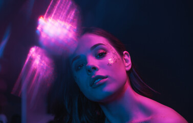 Fashion photo with glare of an attractive woman with bright makeup with glitter on her face, looking at the camera and posing, purple, blue light.