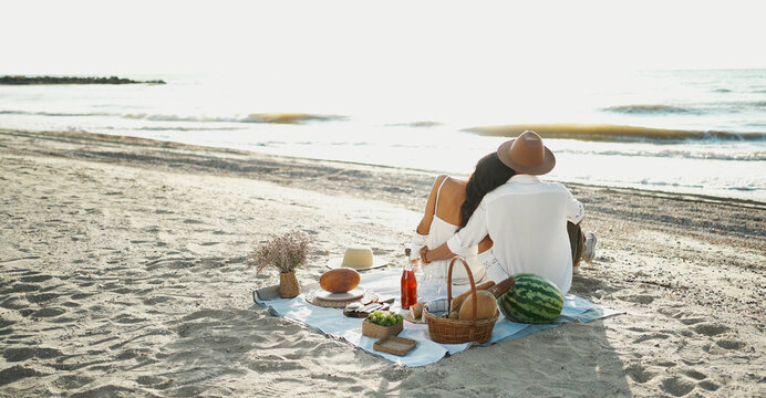 Rear view couple having picnic at the beach, sitting on blanket and embracing, looking to seaview