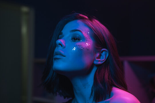 Close photo of a beautiful woman with bright makeup at a night party with purple and blue light, looking away with a serious face.