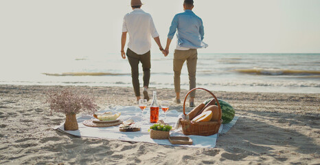 Silhouette gay couple walking by sea beach, focus on picnic blanket with wine, glasses and food....