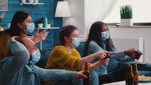 Excited funny young friends with masks holding controllers playing video game at home during new normal party. Overjoyed man and woman winning videogame having fun together on sofa celebrating victory