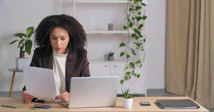Portrait of young professional afro american woman wearing formal suit sitting by desk in home office looking at documents analyzes data checks papers typing on laptop computer, work during lock down
