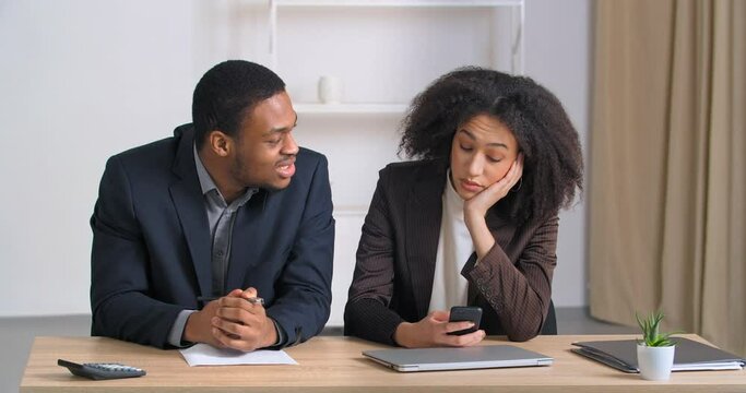 Two colleagues sitting at business meeting ethnic woman manager bored looks at phone chatting online on social networks afro american man snaps his fingers needs attention calling female coworker