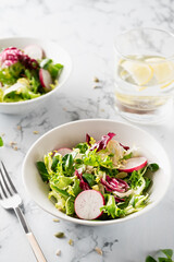 Mesclun with sliced radish in a white bowl with a glass of water and metal fork on a marble surface
