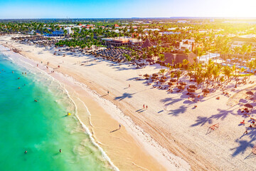 Beach vacation and travel background. Aerial drone view of beautiful atlantic tropical beach with straw umbrellas, palms and boats. Bavaro beach, Punta Cana, Dominican Republic. - obrazy, fototapety, plakaty