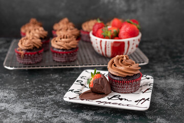 Chocolate Cupcake on Bon Appetit Plate with a Chocolate-Covered Strawberry Against a Black Textured Background; More Chocolate Cupcakes on Cooling Rack in Background with a Bowl of Fresh Strawberries