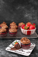Chocolate Cupcake on Bon Appetit Plate with a Chocolate-Covered Strawberry Against a Black Textured Background; More Chocolate Cupcakes on Cooling Rack in Background with a Bowl of Fresh Strawberries