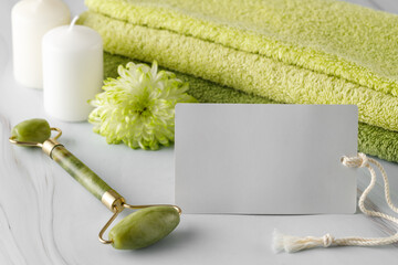 Blank spa  skincare card with jade facial roller for anti-aging skin massage, flower and soft towels on a marble table.