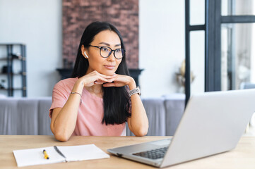 Focused beautiful asian staring on the laptop screen sitting at the desk in the modern home office, watching webinar, reading and recourching